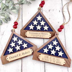May include: Three wooden ornaments shaped like folded American flags with personalized nameplates. The top ornament reads "Veronica Hines, United States Air Force, 3.17.63-7.25.19". The middle ornament reads "Daniel E. Baker". The bottom ornament reads "Home of the Free Because of the Brave".