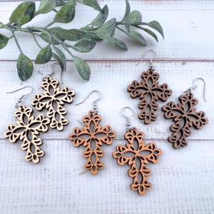 Peut inclure: Boucles d'oreilles en bois en forme de croix, de différentes nuances de brun. Les boucles d'oreilles présentent un motif ajouré détaillé. Les crochets sont de couleur argentée. Elles sont présentées sur une surface en bois blanc avec des brins de feuilles vertes en arrière-plan.