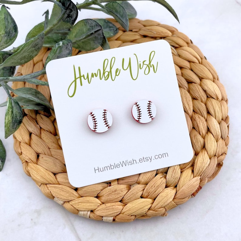 Peut inclure: Boucles d'oreilles &agrave; clous en forme de balles de baseball sur une carte blanche avec le texte "Humble Wish" en vert. Les boucles d'oreilles sont blanches avec des accents rouges et des coutures noires. La carte repose sur une surface tress&eacute;e brun clair.