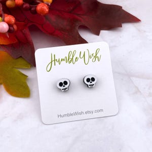 Peut inclure: Une paire de boucles d'oreilles clous en forme de crâne blanc avec des détails noirs, présentées sur une carte blanche. La carte porte l'inscription "Humble Wish" en vert et une adresse de site web. Accessoire sur le thème d'Halloween ou gothique.
