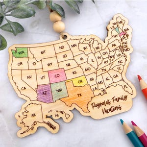 Peut inclure: Ornement en bois en forme de carte des États-Unis, avec des états colorés et le texte "Reynolds Family Vacations". L'ornement est suspendu à une ficelle naturelle et des perles en bois. Des crayons de couleur sont visibles en arrière-plan.