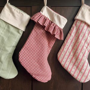 Puede incluir: Tres calcetines navideños de tela colgados en fila. El de la izquierda es verde salvia con finas rayas verticales blancas. El central presenta un patrón de cuadros vichy rojos y blancos con un puño con volantes. El de la derecha tiene rayas verticales rosas y blancas con pequeños detalles florales. Cada calcetín tiene un puño de color crema liso en la parte superior.
