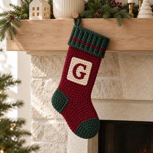 Puede incluir: Un calcetín navideño de ganchillo en hilo rojo oscuro y verde bosque. El calcetín presenta un puño acanalado verde con una fina raya roja, talón y puntera verde oscuro, y un parche cuadrado color crema con una letra G en rojo oscuro.