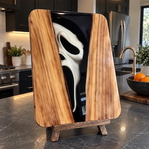 Op de afbeelding: Een rechthoekige houten serveerplank met een centrale verticale inleg van zwarte hars. In de zwarte hars is een wit, langwerpig, vervormd gezichtsmasker met holle ogen en een open mond ingebed, dat doet denken aan een klassiek horrorfilmpersonage.