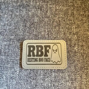 May include: A rectangular light gray patch with rounded corners featuring a dark gray graphic of a ghost with an angry expression. The text RBF is printed in large bold letters above the text RESTING BOO FACE, all enclosed in a dark gray border.