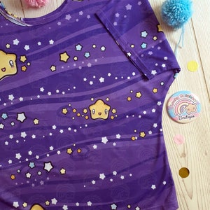 May include: Purple short-sleeved shirt with a repeating pattern of yellow stars, white stars, and blue stars on a purple background with white stripes. The shirt has a smiling yellow star with a face on it. The shirt is on a wooden surface with a small button that says "Lucky Rainbow Boutique" with a rainbow and a star.