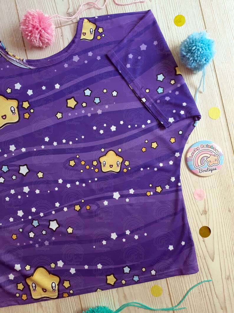 May include: Purple short-sleeved shirt with a repeating pattern of yellow stars, white stars, and blue stars on a purple background with white stripes. The shirt has a smiling yellow star with a face on it. The shirt is on a wooden surface with a small button that says "Lucky Rainbow Boutique" with a rainbow and a star.