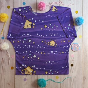 May include: Purple short-sleeved shirt with a galaxy print featuring yellow stars and a cartoon star with a smiling face. The shirt has a relaxed fit and a scoop neck.