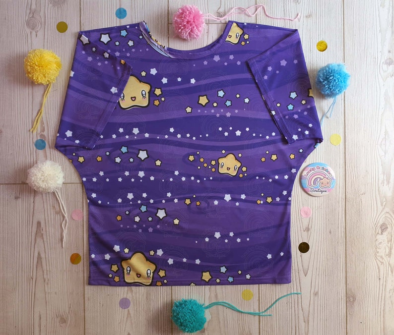 May include: Purple short-sleeved shirt with a galaxy print featuring yellow stars and a cartoon star with a smiling face. The shirt has a relaxed fit and a scoop neck.