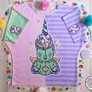 May include: A pink and purple t-shirt with a green and pink zombie unicorn graphic. The shirt has a striped pattern on one side and a swirl pattern on the other. The unicorn has a pink bow and a bone in its hair. The shirt says "Lucky Rainbow Boutique" on the bottom.