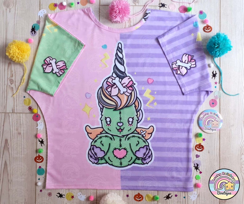 May include: A pink and purple t-shirt with a green and pink zombie unicorn graphic. The shirt has a striped pattern on one side and a swirl pattern on the other. The unicorn has a pink bow and a bone in its hair. The shirt says "Lucky Rainbow Boutique" on the bottom.