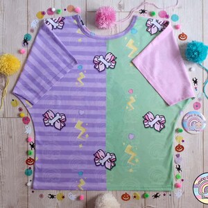 May include: A purple and green striped t-shirt with a pink bow and lightning bolt design. The shirt has a pink short sleeve on one side and a purple short sleeve on the other side.