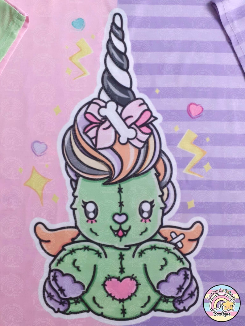May include: A green stitched plush toy with a pink heart, wings, and a unicorn horn. The toy is sitting on a pink and purple striped background with stars and hearts.