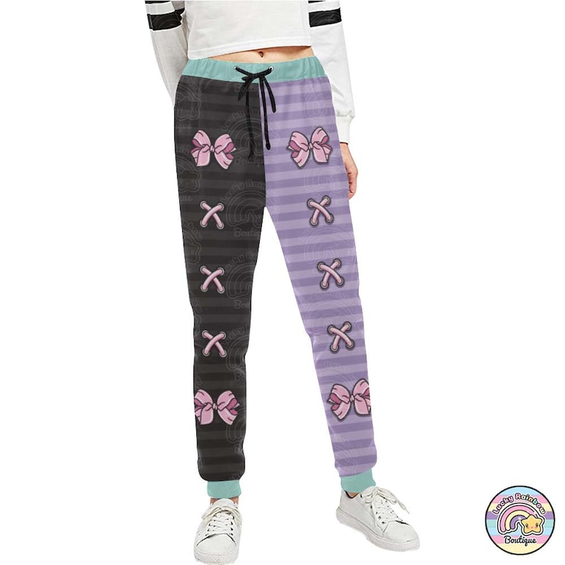 Op de afbeelding: Zwarte en paarse gestreepte sweatpants met roze strikken en een X-patroon. De sweatpants hebben een trekkoord in de taille en een ontspannen pasvorm.