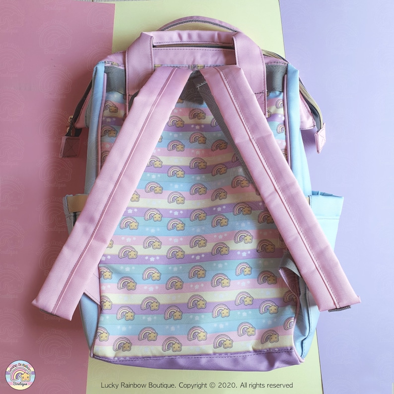 Peut inclure: Un sac &agrave; dos bleu clair avec des bretelles r&eacute;glables roses. Le sac &agrave; dos a un motif de rainbow pastel et blanc avec des &eacute;toiles. Le texte "Lucky Rainbow Boutique. Copyright &copy; 2020. All rights reserved" est imprim&eacute; au bas du sac &agrave; dos.