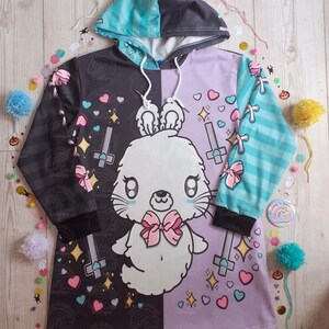 May include: A black and purple hooded sweatshirt with a white bunny rabbit design. The bunny has a pink bow and is surrounded by hearts, stars, and crosses. The sweatshirt has a split design with one side black and the other side purple.