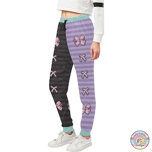 Op de afbeelding: Zwart en paars gestreepte sweatpants met roze strikken en een kruispatroon. De sweatpants hebben een trekkoord.