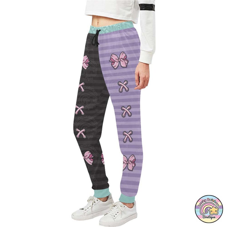 Op de afbeelding: Zwart en paars gestreepte sweatpants met roze strikken en een kruispatroon. De sweatpants hebben een trekkoord.