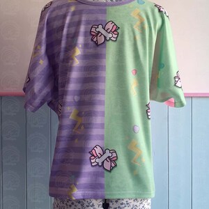 May include: Purple and green t-shirt with a striped pattern on one side and a lightning bolt and bow pattern on the other side. The shirt is on a mannequin.