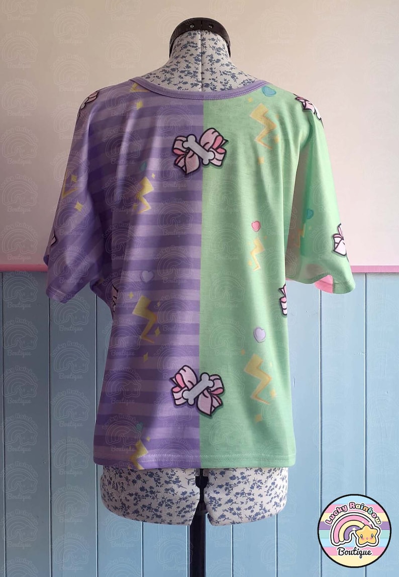 May include: Purple and green t-shirt with a striped pattern on one side and a lightning bolt and bow pattern on the other side. The shirt is on a mannequin.