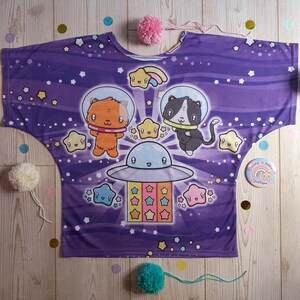 May include: Purple shirt with a space theme featuring a cat and a fox in spaceships with stars and a rainbow. The shirt has a loose fit and a round neckline.