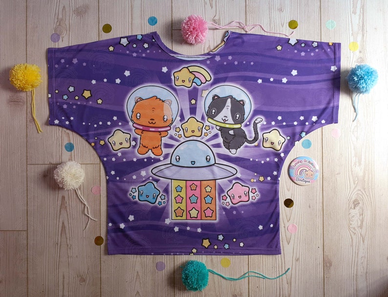 May include: Purple shirt with a space theme featuring a cat and a fox in spaceships with stars and a rainbow. The shirt has a loose fit and a round neckline.