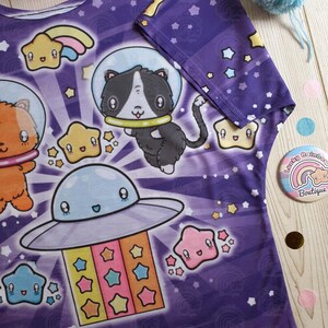 May include: Purple t-shirt with a space-themed print featuring cartoon cats, stars, and a UFO. The print is in a bright, colorful style.