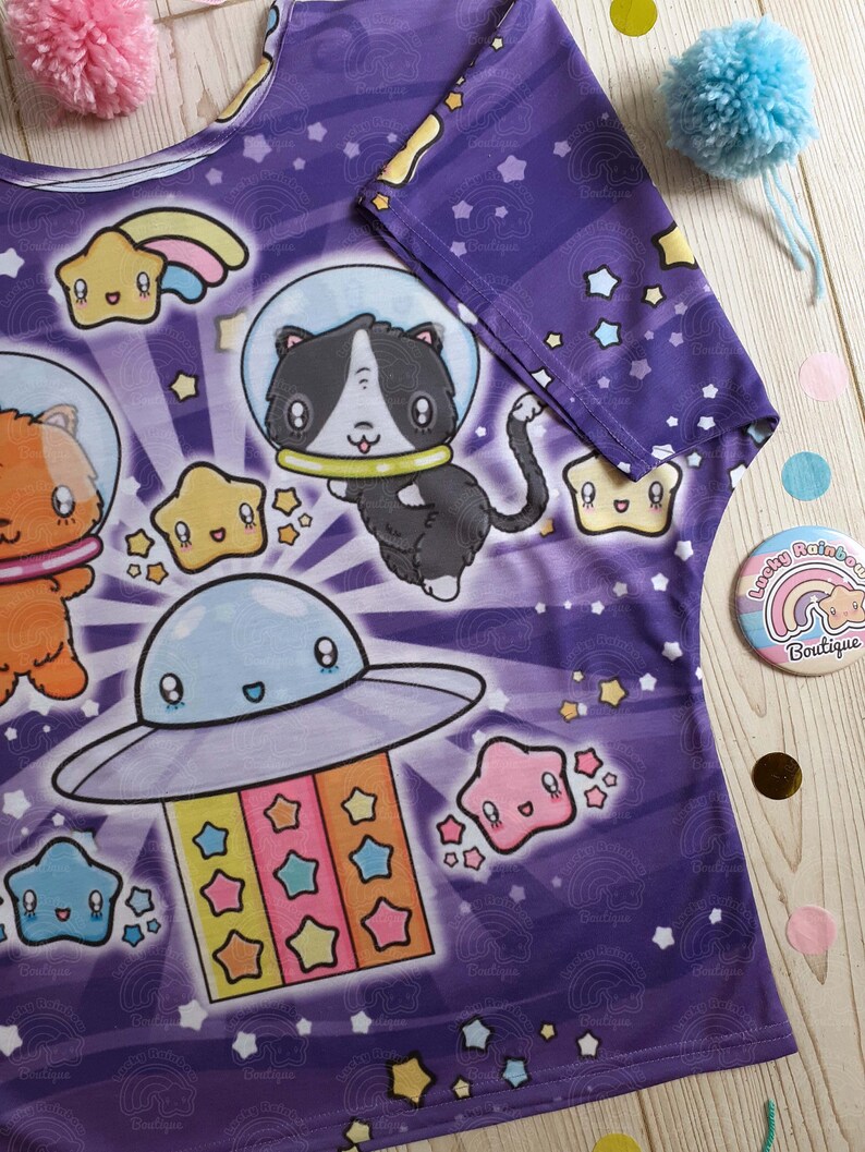 May include: Purple t-shirt with a space-themed print featuring cartoon cats, stars, and a UFO. The print is in a bright, colorful style.
