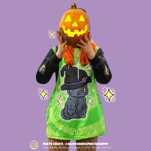 May include: A green and black hooded sweatshirt featuring a cartoon black cat wearing a witch's hat. The cat is surrounded by stars and moons. The model is holding a lit jack-o'-lantern in front of their face.