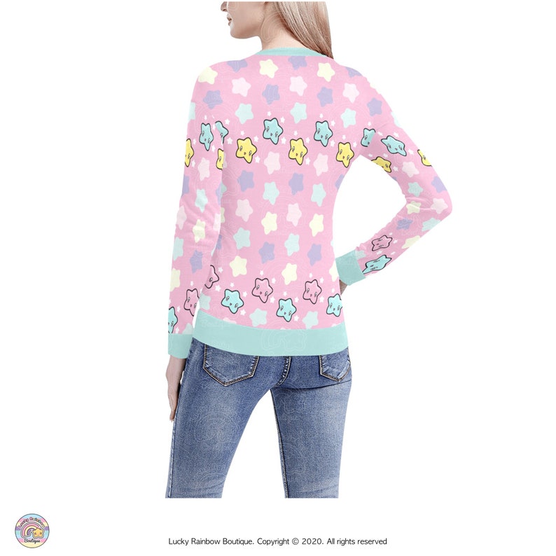May include: A pink long-sleeved sweater with a pattern of pastel-colored stars with faces. The sweater has a light blue ribbed trim around the neck, cuffs, and hem.
