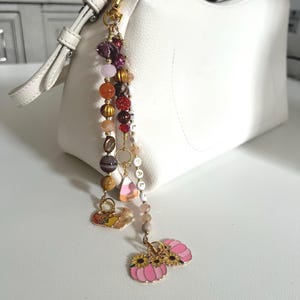 May include: A white handbag featuring a decorative beaded charm attached to the strap. The charm consists of multi-colored beads in shades of pink, orange, brown, and gold, interspersed with small metal charms including pink pumpkins and sunflowers.
