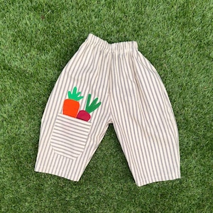 May include: A pair of off-white trousers featuring thin vertical dark stripes and an elasticized waistband. A patch pocket on the front displays two fabric vegetable appliqués, specifically an orange carrot and a purple beet with green tops.