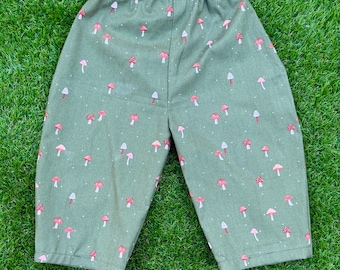 May include: Olive green pants featuring an elasticized waistband and a repeating pattern of small red and white mushrooms scattered across the fabric. The garment has a straight-leg cut and a simple hem at the bottom of each leg opening.