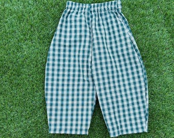 May include: A pair of trousers featuring a dark green and white plaid pattern. The garment has an elasticated waistband and a relaxed, wide-leg silhouette that tapers slightly towards the hem, constructed from a woven fabric.