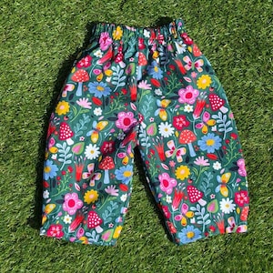 Puede incluir: Un pantalón con cintura elástica sobre un fondo verde oscuro estampado con un patrón colorido de setas rojas y blancas, diversas flores azules, rosas, amarillas y blancas, hojas verdes y pequeñas mariposas amarillas.
