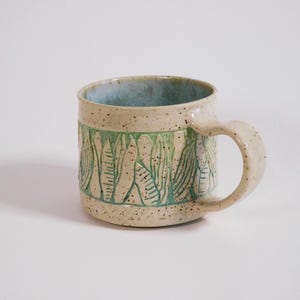 May include: A ceramic mug with a speckled cream glaze and a light blue interior. The exterior features a central band of carved, vertical leaf-like patterns highlighted in green. The mug has a sturdy, curved handle and a cylindrical body shape.