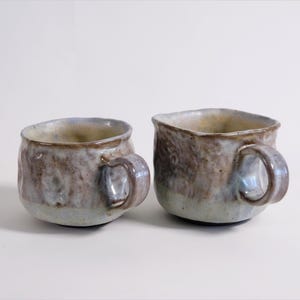Peut inclure: Deux tasses en céramique faites à la main avec une surface texturée et irrégulière. L'émail présente un mélange de tons brun terreux, bleu sourd et blanc crème. Chaque tasse a un corps arrondi, une large ouverture et une petite anse incurvée.