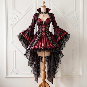 Pode incluir: Um vestido vermelho escuro e preto com bainha assimétrica, mangas longas e largas com acabamento em renda, corpete estilo espartilho com bordados decorativos e gola alta. A peça inclui uma gargantilha de renda preta com um pingente vermelho.