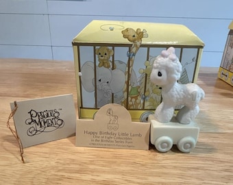 Może przedstawiać: Biała porcelanowa figurka jagnięcia z różową kokardką, stojąca na podstawie w kształcie wagonu kolejowego z numerem jeden. Zestaw zawiera żółte pudełko z ilustracjami zwierząt z zoo oraz etykietę z napisem Precious Moments Happy Birthday Little Lamb.