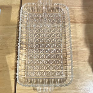 May include: A clear glass rectangular serving tray featuring a geometric diamond pattern across the center and a scalloped edge border. The tray includes a small divided section at the top and two integrated handles on the short ends.
