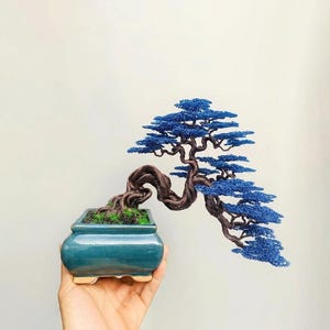 May include: A wire bonsai tree sculpture featuring a twisted dark brown trunk and branches with vibrant blue foliage. The tree is planted in a square, glossy teal ceramic pot with a small amount of green moss at the base of the trunk.