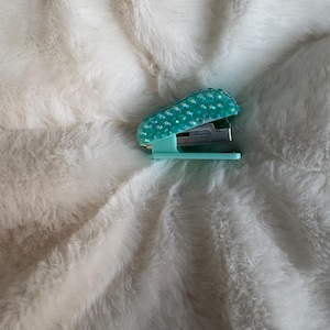 May include: A small mint green stapler featuring a top surface covered in iridescent, light-reflecting rhinestones. The base and handle are a solid mint green plastic, while the internal stapling mechanism is made of polished silver metal.