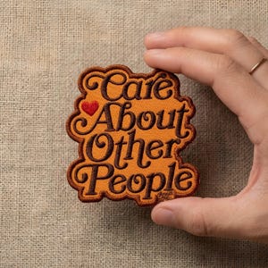May include: An embroidered patch with an orange background and dark brown cursive lettering that reads Care About Other People. A small red heart is positioned next to the word Care. The patch has a scalloped edge and a dark brown border.