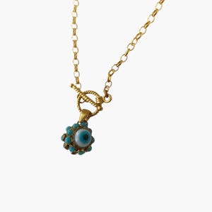 May include: Gold-toned necklace featuring a pendant with a blue and white eye design, surrounded by turquoise-colored beads. The necklace has a toggle clasp and a delicate chain.