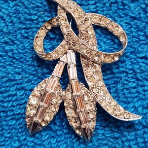 May include: A silver-toned brooch featuring a ribbon-like design adorned with numerous small round clear rhinestones and three dangling leaf-shaped pendants. Each leaf pendant is set with a central row of rectangular baguette-cut clear rhinestones.