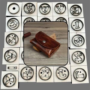 May include: A brown leather card holder with a snap button closure is centered over a collection of square wooden blocks, each featuring a circular metal die cutter shaped like a different letter of the alphabet.