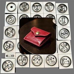 May include: A small red leather coin purse with a gold-tone metal snap button closure and white stitching, centered over a background of multiple square wooden blocks, each featuring a unique metal letter-shaped cutting die.