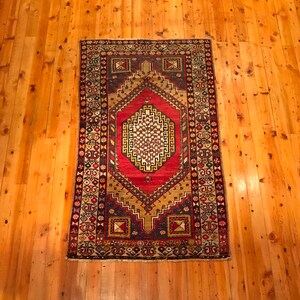 May include: A rectangular rug with a central red diamond design and a gold and black border. The rug has intricate geometric patterns in red, gold, blue, and white. It is placed on a wooden floor.
