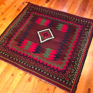 May include: A square, hand-woven rug with a geometric design. The rug features a deep burgundy field with a central diamond and zig-zag patterns in red, green, and navy blue. The border is a repeating floral pattern in black and white.