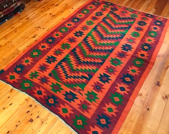 May include: A rectangular, hand-woven rug with a geometric pattern. The rug features a central zig-zag design in shades of green, orange, and burgundy, framed by borders of repeating star-like shapes in blue and orange. The rug is displayed on a wooden floor.